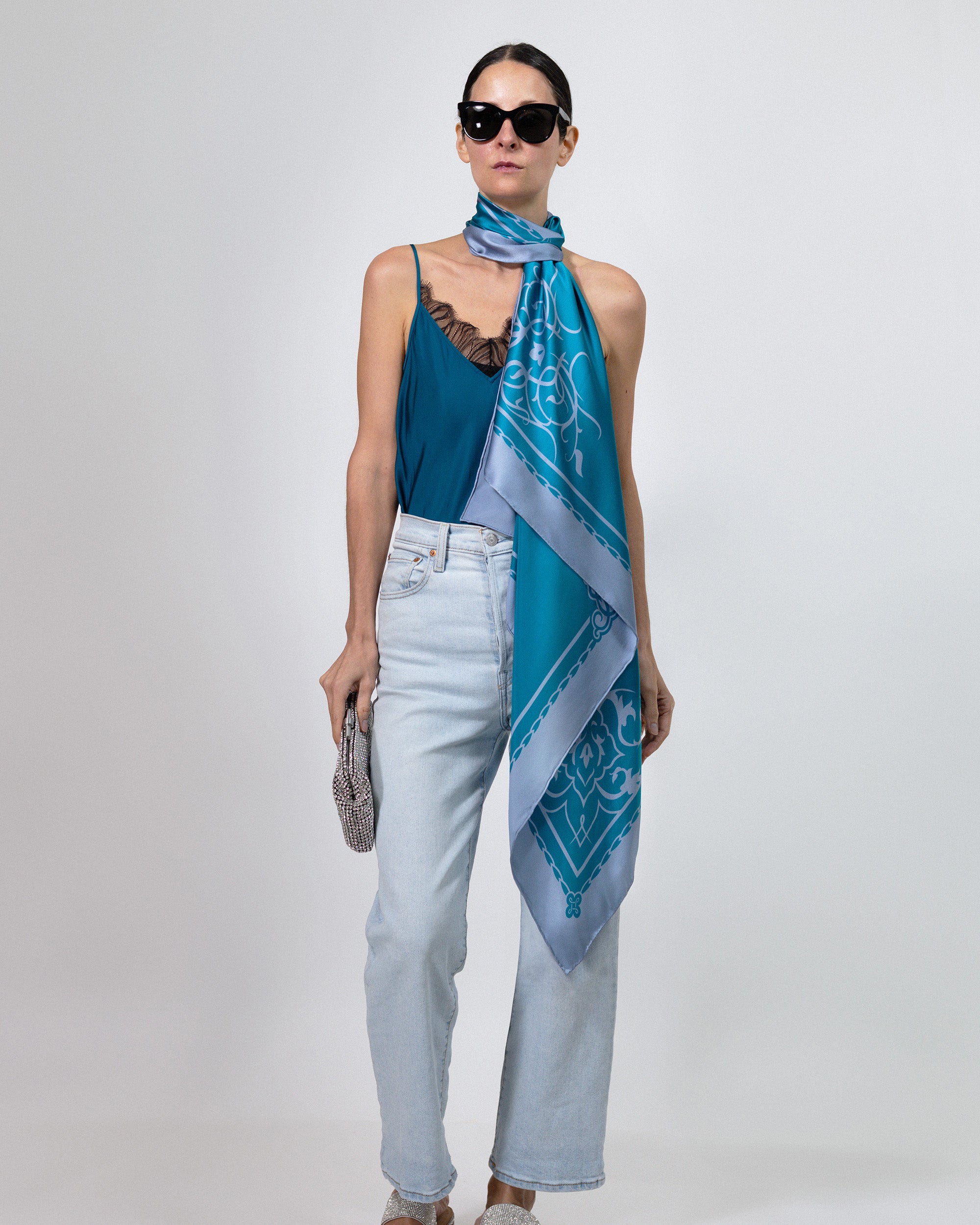 A model wearing a blue and teal patterned silk scarf wrapped around her shoulders, styled with a teal top, light-wash jeans, and sunglasses against a light gray studio background.