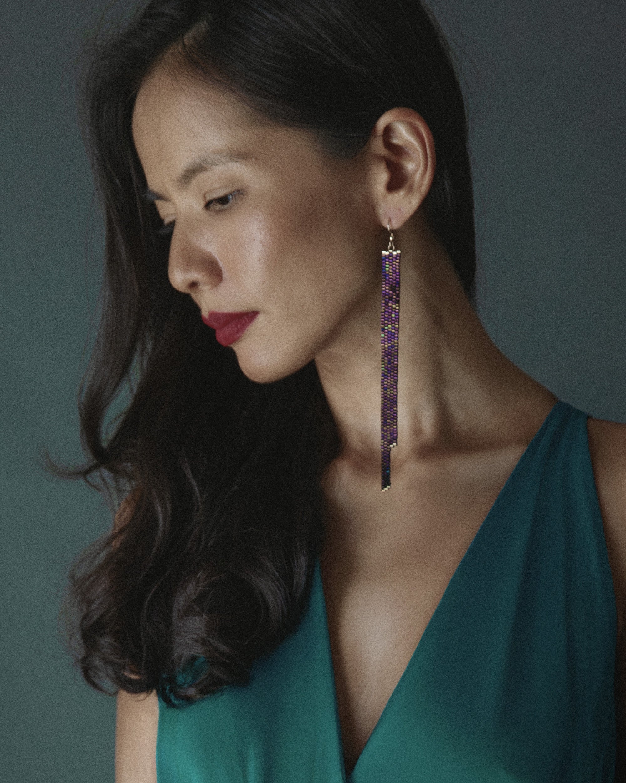 Woman wearing a green dress with a deep neckline, dark hair, and purple earrings against a gray background.