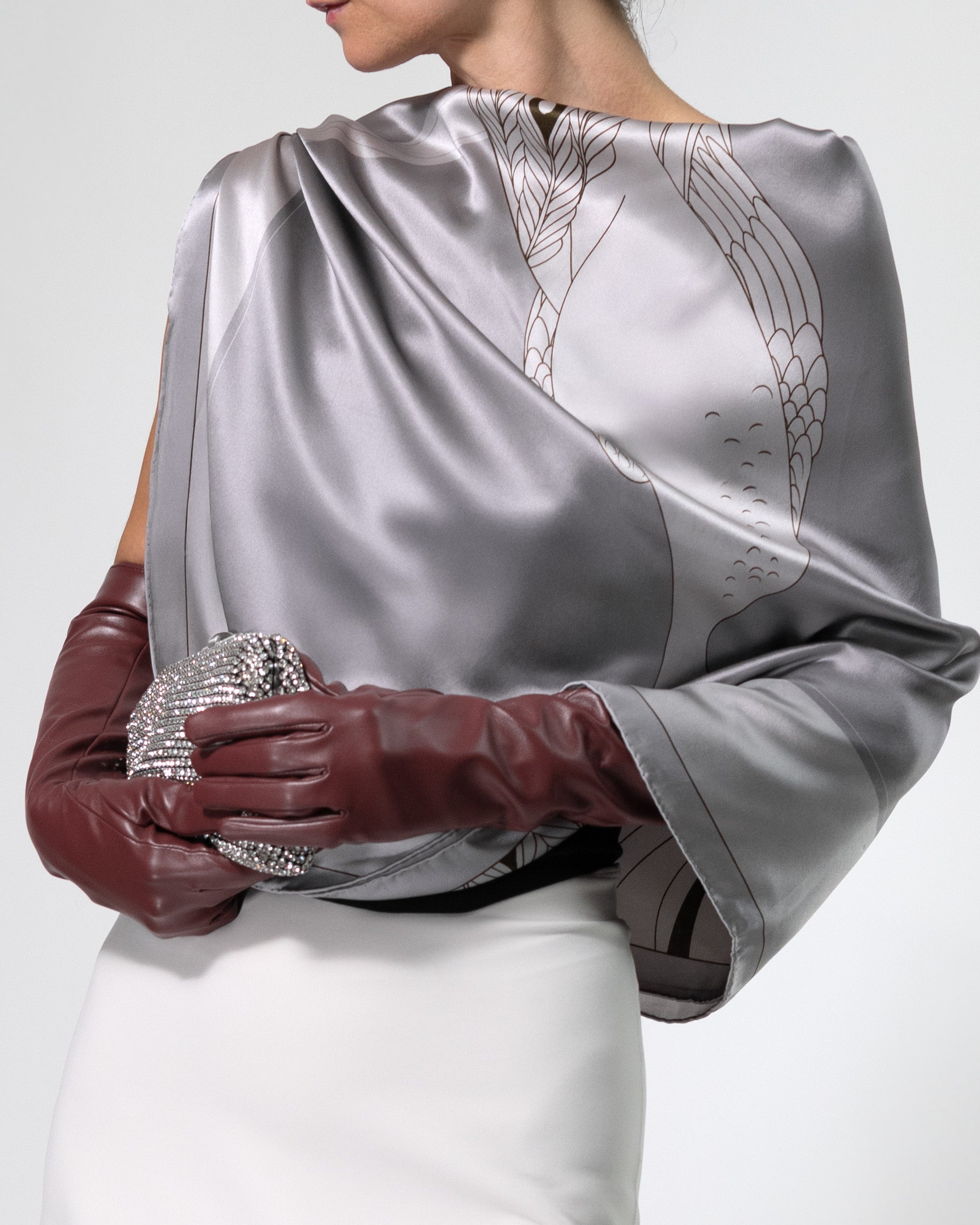 Person wearing a silver satin scarf with peacock feather design, white dress, and burgundy gloves holding a clutch.