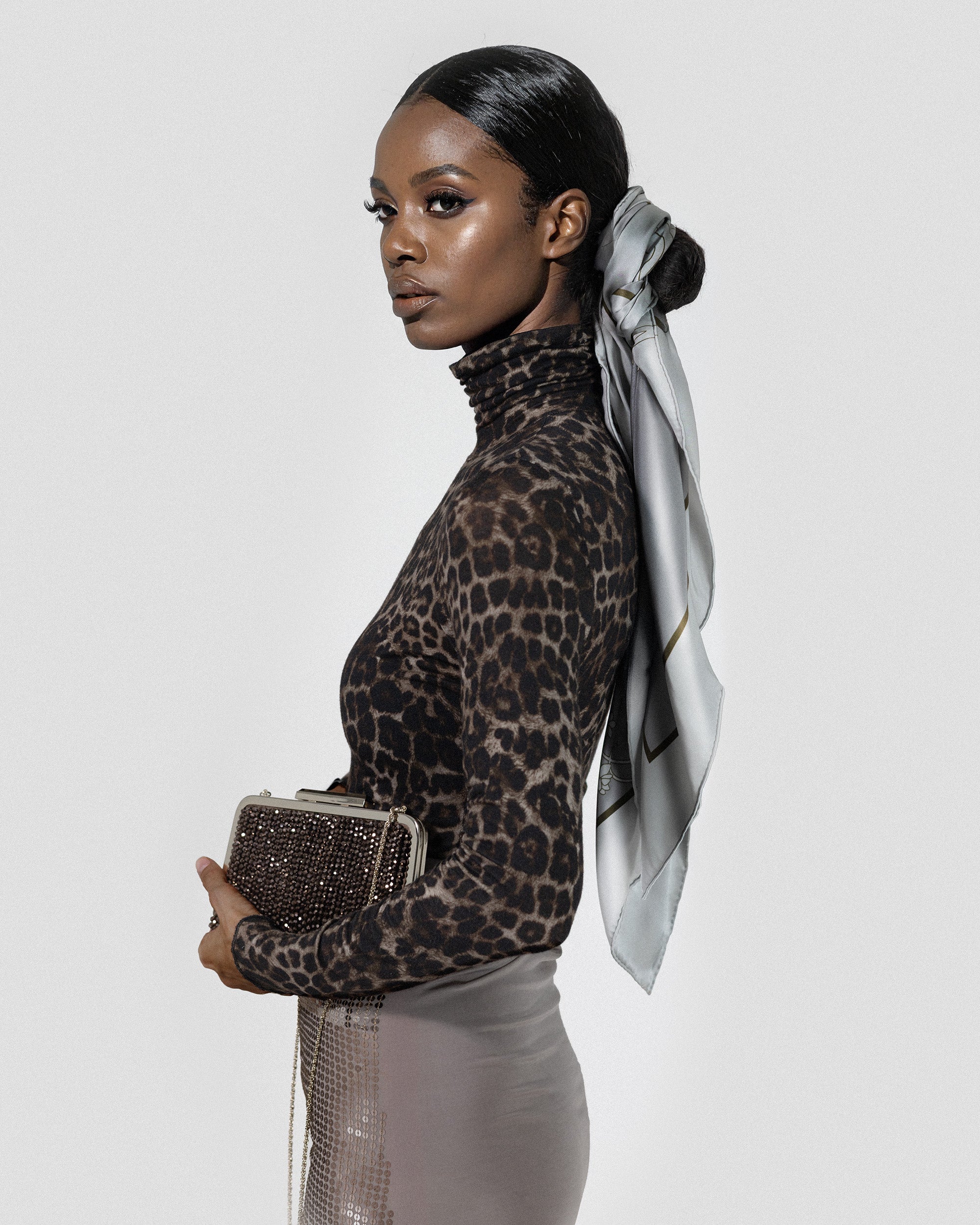 Woman wearing a leopard print top, silk scarf wrapped around hair knot bun, and holding a clutch with a white background