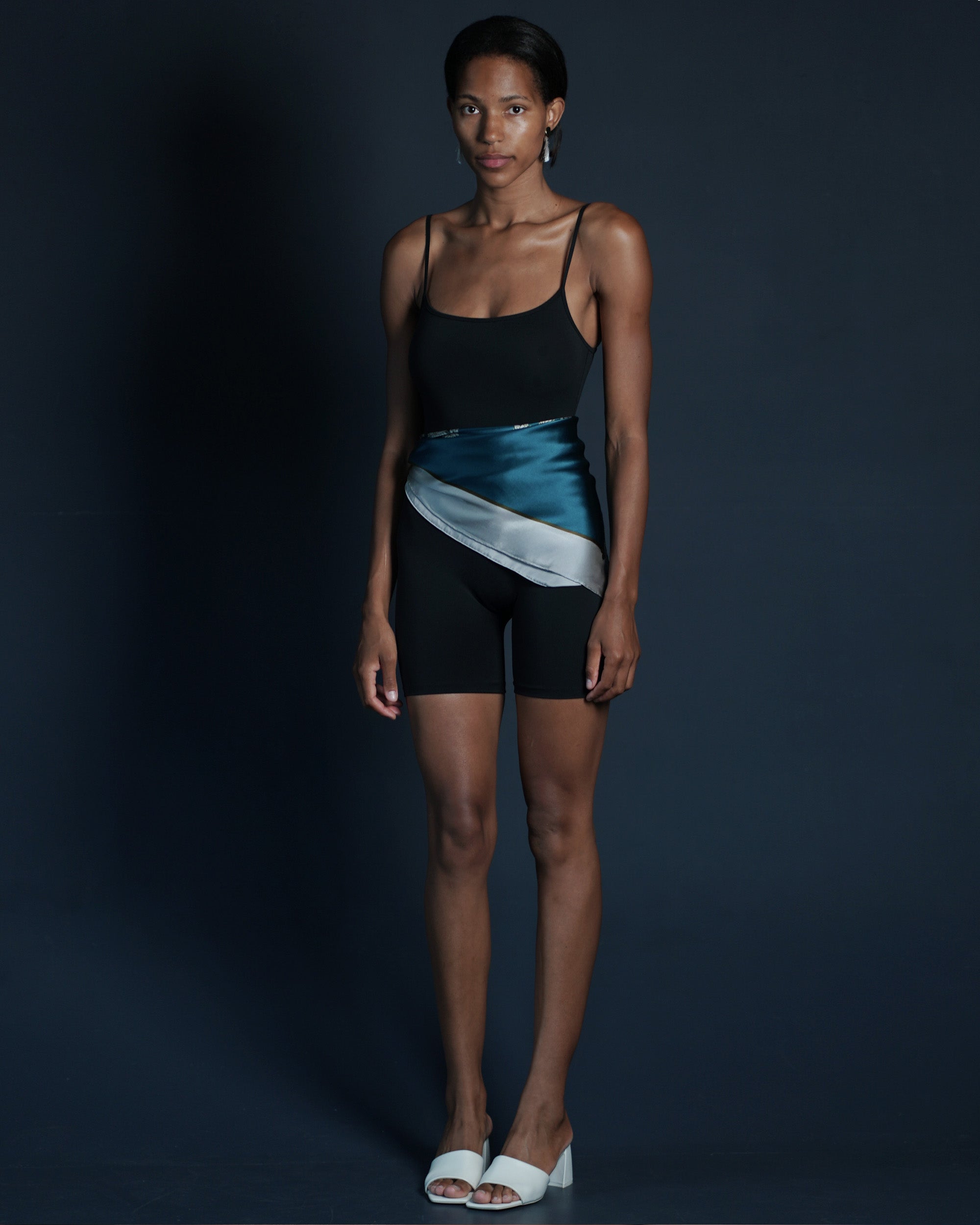 Model wearing a black sport jumpsuit with a silk scarf around waist against a dark background