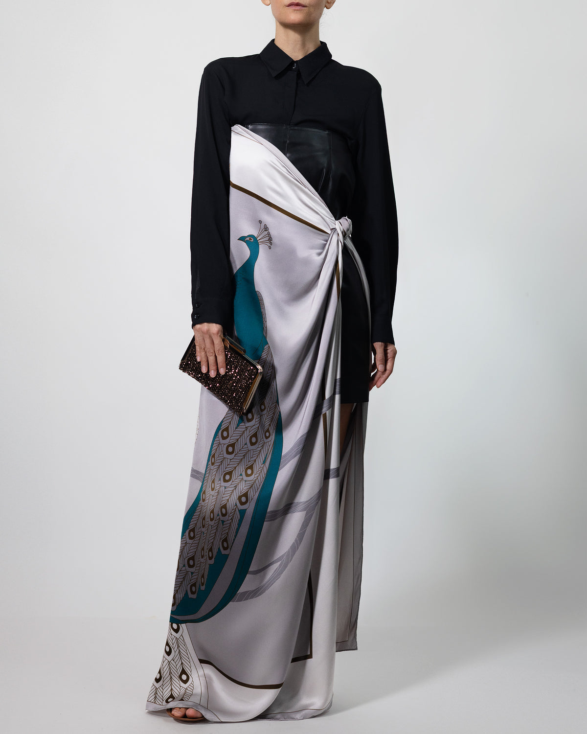 A model wearing a long-sleeved black collared shirt and a floor-length white silk scarf HELGA-DESIGN brand with a printed peacock design, holding a small brown clutch against a light grey studio background.