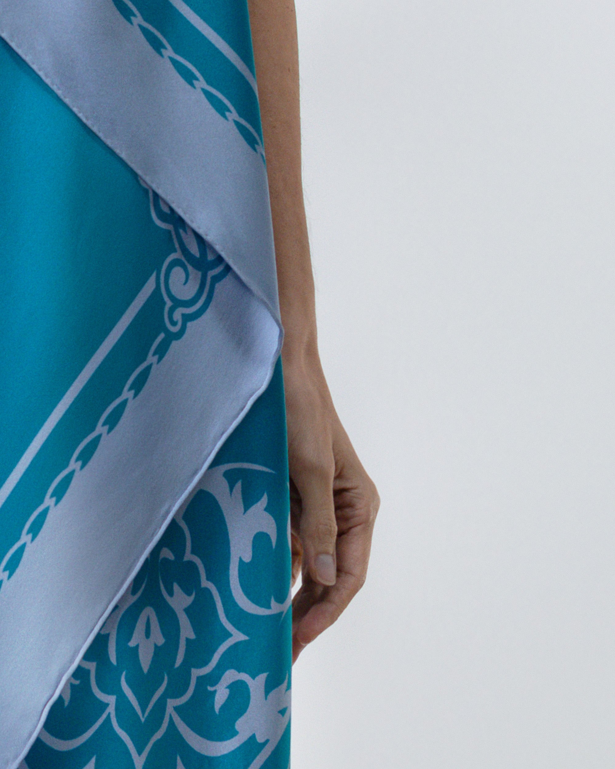 A close-up of a person holding a turquoise and light blue patterned scarf or shawl against a plain light gray background.