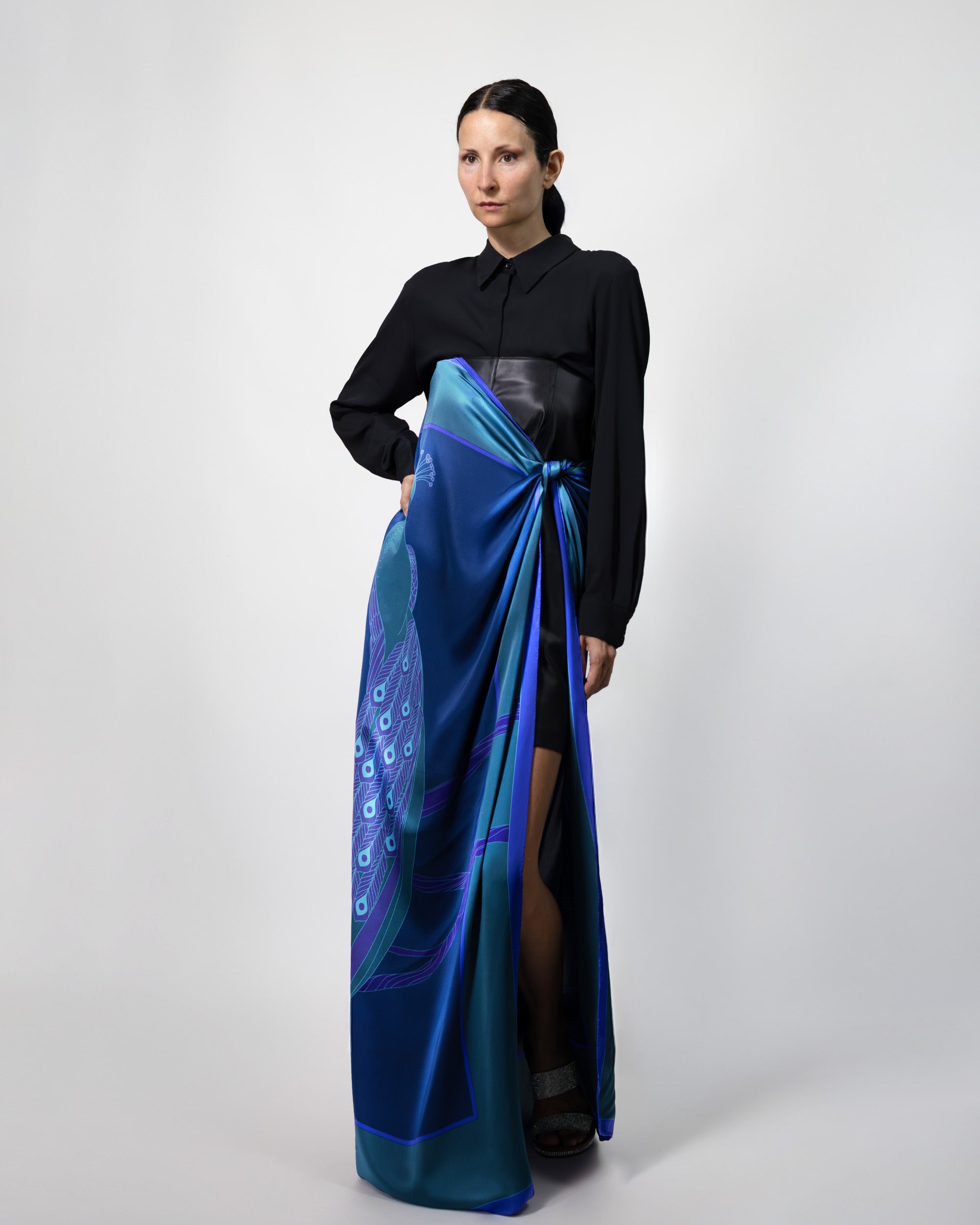 A female model wearing a black outfit with a deep blue and green silk scarf on a white background